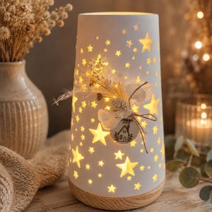 Könnte beinhalten: Eine zylindrische weiße Keramiklampe mit zahlreichen sternförmigen Ausschnitten, die ein warmes Licht abgeben. Der Sockel besteht aus hellem Holz. Ein kleiner Strauß getrockneter Blumen und ein rundes Holzschild mit einer Hauszeichnung sind vorne befestigt.