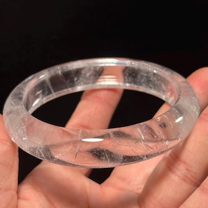 Puede incluir: Un brazalete de cuarzo transparente. El brazalete transparente es circular y tiene una superficie lisa y pulida. El cuarzo tiene inclusiones e imperfecciones naturales, lo que se suma a su carácter único. El fondo es oscuro.
