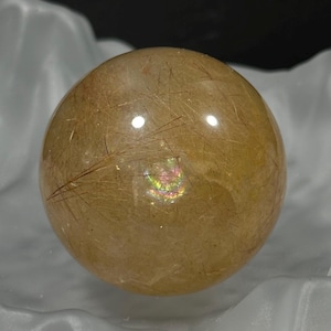 May include: A golden-brown crystal ball with internal inclusions and a polished surface. The sphere has a reflective quality, with light refracting within, creating a rainbow effect. The crystal ball is a decorative item.