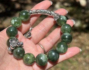 May include: A green jade stone bracelet with silver accents. The bracelet features a floral design and a clasp with a dangling charm.