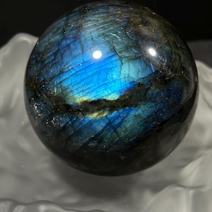 Può includere: Una sfera di labradorite lucidata, che mostra un affascinante gioco di colori. La superficie dell'orbita presenta iridescenze blu, verdi e sfumature dorate. La gemma è posta su uno sfondo bianco morbido, che ne esalta la bellezza naturale.