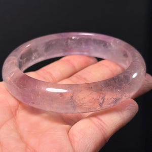 Puede incluir: Un brazalete de cuarzo translúcido de color púrpura claro. El brazalete liso y redondeado se sostiene en una mano sobre un fondo negro. El color del brazalete varía de transparente a un suave tono lavanda, con inclusiones naturales visibles.