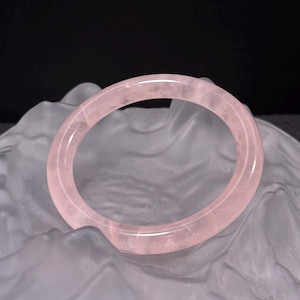 Könnte beinhalten: Ein durchscheinendes, hellrosa Rosenquarz-Armreif-Armband. Das glatte, kreisförmige Armband wird vor einem weichen, strukturierten Hintergrund präsentiert. Der Rosenquarz hat eine blassrosa Farbe.