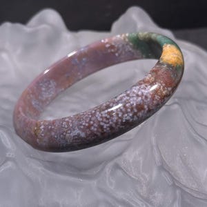 May include: A round gemstone bangle. The bracelet is a mix of colours, including red, green, yellow, and white. The surface has a speckled pattern. The bracelet is displayed on a white, textured surface.