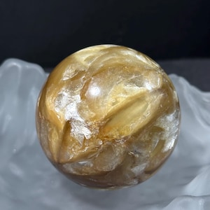 Könnte beinhalten: Eine goldfarbene Kugel mit einem wirbelnden, strukturierten Muster. Die Kugel liegt auf einer weißen, wellenförmigen Oberfläche vor einem dunklen Hintergrund. Die Kugel scheint durchscheinend zu sein, wobei sich das Licht auf ihrer Oberfläche spiegelt. Durchmesser ca. 7 cm.