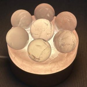 May include: A white crystal lamp base with seven clear quartz crystal spheres on top. The base is lit from within, illuminating the crystals.