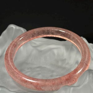 Op de afbeelding: Een doorschijnende, rozerode edelsteenarmband. De cirkelvormige armband is glad en gepolijst en toont natuurlijke insluitsels. De armband wordt gepresenteerd op een wit, getextureerd oppervlak tegen een donkere achtergrond. Een sieraad.
