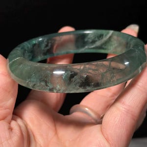 Op de afbeelding: Een doorschijnende groene jade armband. De gepolijste steen heeft een glad oppervlak met interne adering en insluitsels, waardoor een uniek patroon ontstaat. De armband wordt in een hand gehouden tegen een donkere achtergrond.