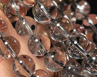 Könnte beinhalten: Eine Kette aus klaren Quarzperlen mit schwarzen Einschlüssen. Die Perlen sind rund und haben eine glatte, polierte Oberfläche.