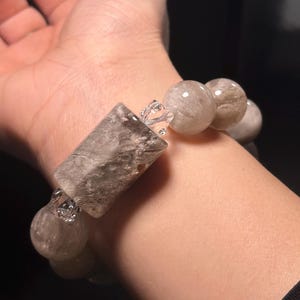 May include: A grey and white crystal bracelet with a rectangular bead. The bracelet is made of round and rectangular beads.