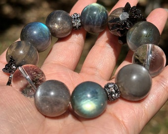 May include: A bracelet made of dark gray and blue gemstone beads with silver accents. The bracelet has a flower-shaped charm with black and silver accents.