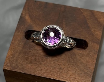 May include: A silver ring with a large, round, faceted purple gemstone. The ring is set in a decorative bezel and rests on a dark wooden block. The ring's band features intricate scrollwork. The background is a blurred, gray surface.
