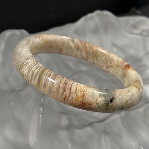 May include: A natural stone bangle bracelet. The bracelet displays a mix of beige, tan, and reddish-brown tones with a striped pattern. The smooth, polished surface reflects light, highlighting its unique colour variations. A beautiful piece of jewellery.