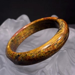 Op de afbeelding: Een gepolijste, cirkelvormige armband. De armband heeft een mix van goud, bruin en rood, met een natuurlijke, getextureerde uitstraling. De sieraden worden gepresenteerd op witte, gedrapeerde stof tegen een donkere achtergrond.