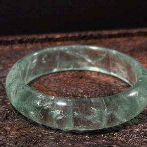 May include: A translucent green jade bangle bracelet. The smooth, rounded bracelet is a solid band of pale green stone. The jade has natural variations in color and texture. The bracelet is displayed on a dark, textured surface.