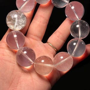 May include: A bracelet of translucent, round beads in shades of pink, clear, and peach. The beads are strung together, forming a circular shape. The bracelet is held against a dark background, highlighting the colours and clarity of the stones.