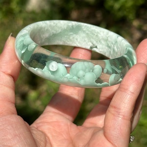 Könnte beinhalten: Ein hellgrünes Harz-Armband mit durchscheinendem, wolkigen Aussehen. Das Armband hat eine glatte, abgerundete Oberfläche und ist mit kleinen, weißen, undurchsichtigen Kreisen verziert.