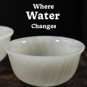 Könnte beinhalten: Eine weiße, durchscheinende Schale mit abgerundeter Form, vor dunklem Hintergrund. Die Schale hat ein marmoriertes Aussehen mit dezenten grauen Adern. Der Text "Where Water Changes" ist weiß über der Schale angezeigt.
