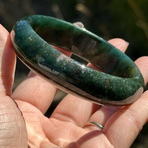 Può includere: Un braccialetto bangle in giada screziata verde e bianca. La pietra liscia e lucida è di un verde foresta intenso con inclusioni verdi e bianche più chiare. Il testo "NO.1" è in basso nell'immagine.