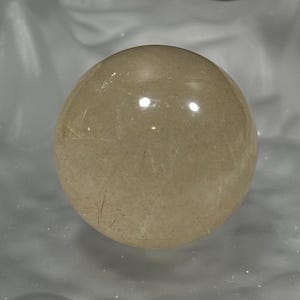 以下が含まれることがあります： 内部にインクルージョンを持つ、半透明の球形の宝石が、質感のある明るい色の表面に置かれています。球体は研磨されており、光を反射して明るいハイライトを作り出しています。背景は暗く、球体の形を際立たせています。