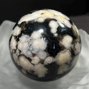 Peut inclure: Une sphère d'agate noire et blanche avec un motif tourbillonnant d'inclusions blanches et rose pâle. La sphère est polie et lisse.