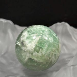 Könnte beinhalten: Eine transparente grüne Fluoritkugel mit weißen Adern. Der polierte Kristallball wird vor einem weißen, strukturierten Hintergrund mit dunklem Hintergrund präsentiert. Ein einzigartiger Dekorationsartikel.