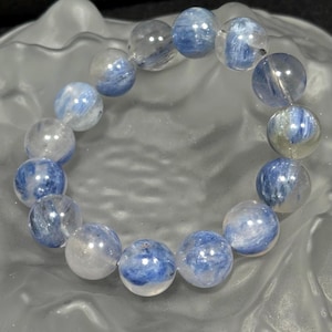 Könnte beinhalten: Ein Armband aus kugelförmigen Perlen. Die Perlen sind eine durchscheinende, helle Farbe mit Wirbeln von Blau. Das Armband liegt auf einer strukturierten, hellfarbigen Oberfläche mit dunklem Hintergrund.