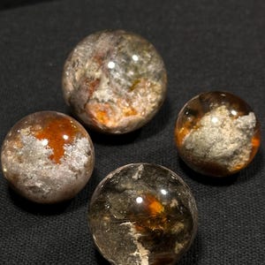 May include: Four clear quartz spheres with inclusions of orange, brown, and white. The spheres are arranged on a dark gray textured surface.