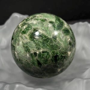 May include: A green sphere with a textured surface, set in a white, wavy dish. The sphere has a mottled appearance with varying shades of green and lighter veins. The dish has a glossy, reflective surface. The sphere is likely made of stone.