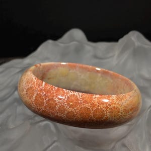 Puede incluir: Un brazalete de color coral con un patrón floral. El brazalete tiene una superficie lisa y pulida y un tono cálido y terroso. El brazalete tiene forma circular y parece estar hecho de un material natural. El fondo es una superficie suave, blanca y texturizada.