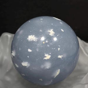 May include: A smooth, blue-grey sphere with white, cloud-like inclusions. The polished stone sits on a reflective, silvery surface, against a dark background. This decorative sphere is a unique home decor item.