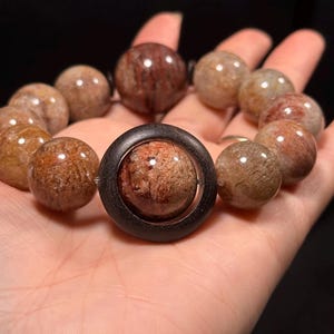 Könnte beinhalten: Ein Armband mit kugelförmigen, rötlich-braunen Edelsteinen mit gesprenkeltem Aussehen. Das Armband enthält eine größere, zentrale Perle, die in einem dunklen Holzring gefasst ist. Die Perlen werden vor einem dunklen Hintergrund präsentiert.