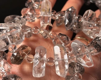 Könnte beinhalten: Eine Kette aus klaren Quarzkristallperlen mit schwarzen Einschlüssen. Die Perlen sind facettiert und haben verschiedene Formen, darunter lange, dünne und runde. Die Perlen sind auf einer durchsichtigen Gummikordel aufgefädelt.