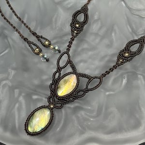 May include: A handcrafted macrame necklace with a pendant featuring an oval, iridescent gemstone. The necklace is brown with intricate knotting and small gold-colored beads. Matching earrings with similar gemstones and macrame details are also present.
