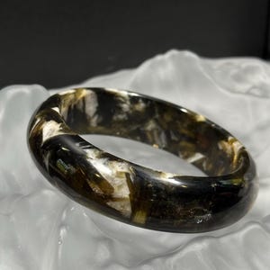 May include: A polished, oval-shaped bracelet with a marbled appearance. The bracelet features a mix of dark green, brown, and white tones, creating a unique pattern. The jewelry is displayed on a white, textured surface.