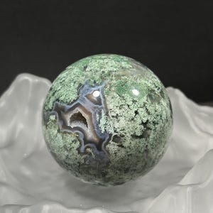 May include: A spherical moss agate gemstone with a green, white, and grey pattern. The polished stone has a geode-like cavity in the centre. It rests in a white, textured, decorative bowl. The background is black.