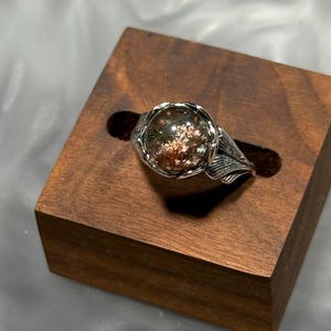Könnte beinhalten: Ein silberner Ring mit einem großen, runden, klaren Stein mit grünen und kupferfarbenen Einschlüssen. Der Ring hat ein blattartiges Design um den Stein und wird auf einem kleinen, dunklen Holzblock präsentiert.