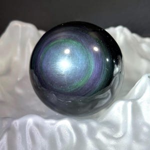 May include: A glossy, spherical gemstone displaying a swirling pattern of iridescent colours, including blue, green, and purple. The orb is set against a backdrop of white fabric, creating a striking contrast. This decorative item is a unique home decor piece.
