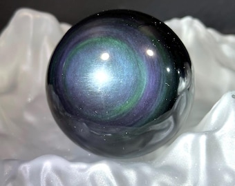 May include: A polished, spherical gemstone with a swirling pattern of iridescent colors, including blue, green, and purple. The orb is set against a backdrop of white fabric, creating a striking contrast. This decorative item is a unique home decor piece.