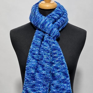 May include: A knitted scarf featuring a variegated pattern of multiple shades of blue, including navy, royal blue, and light blue. The scarf has a textured, chunky knit construction and is draped around the neck of a black display mannequin.