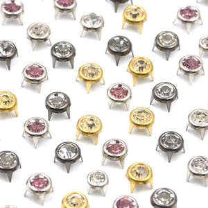 Op de afbeelding: Assortiment van ronde, decoratieve metalen studs met heldere, roze en gouden strass steentjes. De studs hebben drie pinnen aan de onderkant om te bevestigen aan stof of andere materialen.