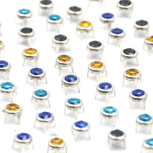 Puede incluir: Surtido de piedras de imitación de colores con engastes plateados. Las piedras de imitación son azules, amarillas y turquesas. Las piedras de imitación están dispuestas en filas sobre un fondo blanco.