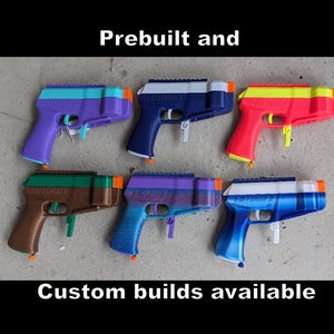 May include: Six colorful toy guns in various color combinations, including purple, blue, red, and brown. The text "Prebuilt and Custom builds available" is displayed above and below the guns.