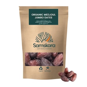Może przedstawiać: Brązowa papierowa torebka zawierająca organiczne daktyle Medjoul w rozmiarze jumbo. Etykieta zawiera napis Organic Medjoul Jumbo Dates oraz nazwę marki Samskara. Przezroczyste okienko ukazuje ciemnobrązowe, pomarszczone daktyle, z kilkoma luźnymi obok.