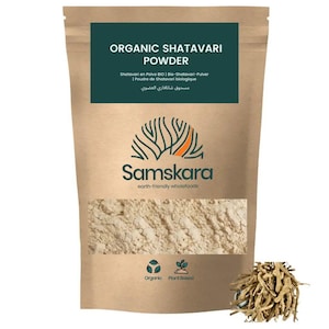 Pode incluir: Uma bolsa de papel castanho contendo pó de shatavari orgânico. O rótulo apresenta o texto Organic Shatavari Powder, o logótipo Samskara e ícones de orgânico e vegetal. À direita, encontra-se um pequeno monte de raízes secas de shatavari.