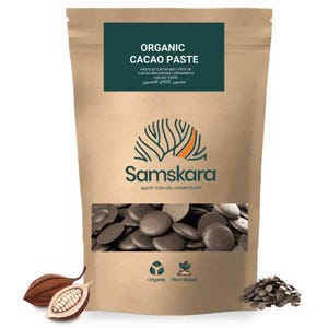 May include: A brown paper pouch of Samskara Organic Cacao Paste featuring a dark green label with white text. The packaging displays a tree logo, organic and plant-based icons, and a clear window showing dark brown cacao discs inside.