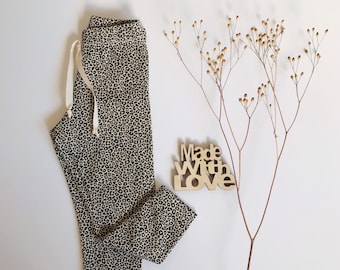 May include: Black and beige leopard print leggings with a white drawstring. The leggings are folded in half and laid flat on a white surface. A wooden sign with the text "Made with Love" is in the background.