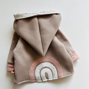 Könnte beinhalten: Taupefarbener Baby-Hoodie mit Kapuze und Regenbogen-Applikation auf dem Rücken. Der Regenbogen hat rosa, weiße und taupefarbene Farben. Die Bündchen und das Kapuzenfutter sind rosa mit einem Tupfenmuster.