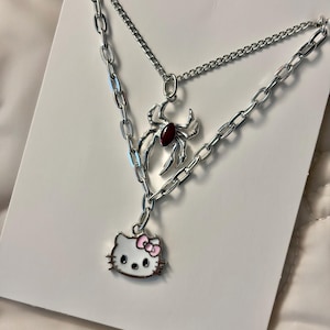 May include: A silver-toned layered necklace featuring a spider pendant with a red gem and a Hello Kitty charm with a pink bow. The necklace has two chains, one with a curb link design and the other with a thinner chain.