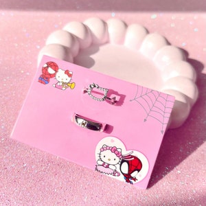 May include: A pink card with a white spiderweb design. The card features two cartoon characters, Hello Kitty and Spiderman, and three silver rings. The rings are a simple band, a ring with a heart design, and a ring with a square design with small stones.